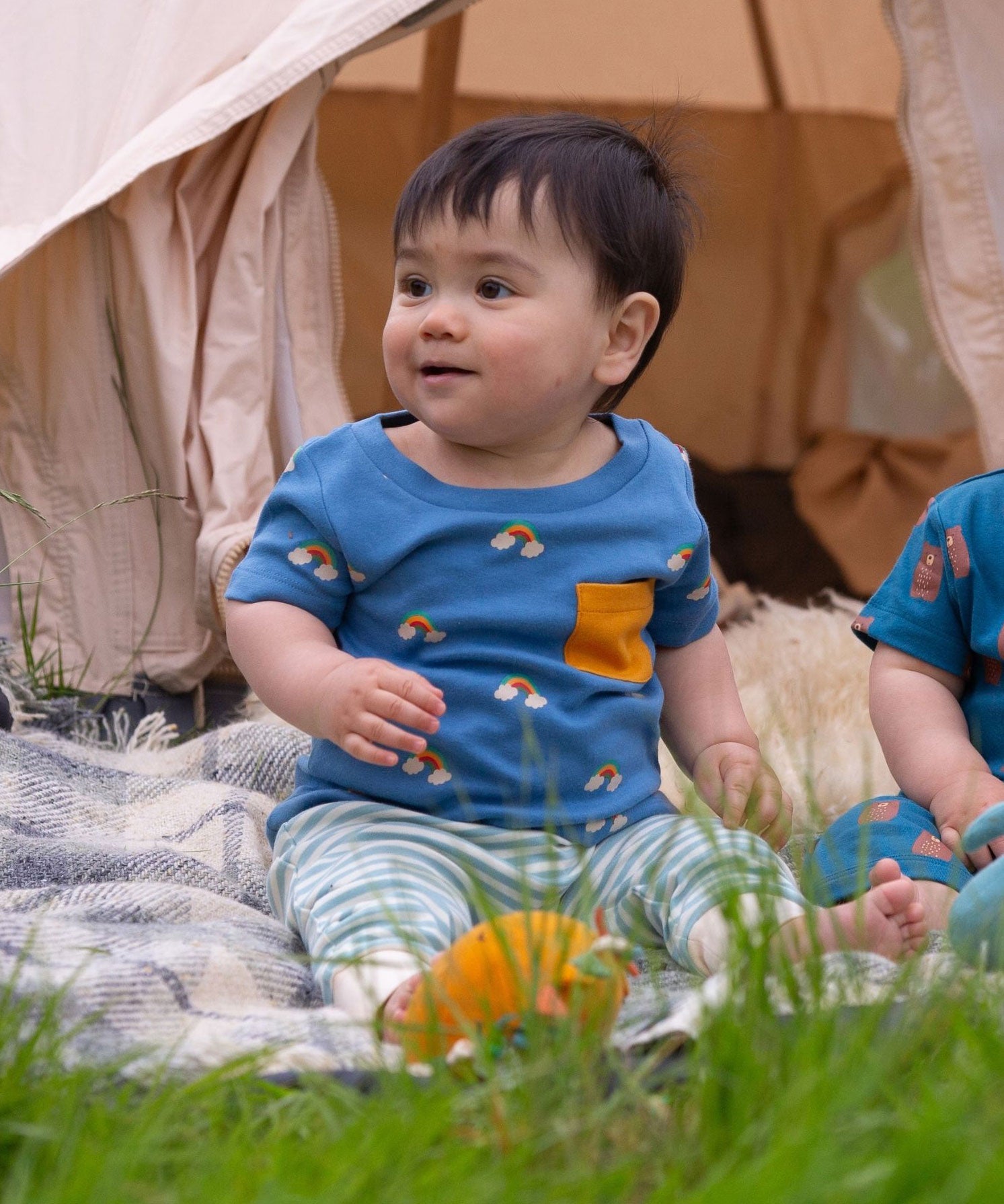 A young child sitting outdoors wearing the Little Green Radicals Kids' Organic T-shirt and Joggers Playset outfit in the  Blue Rainbows design. 