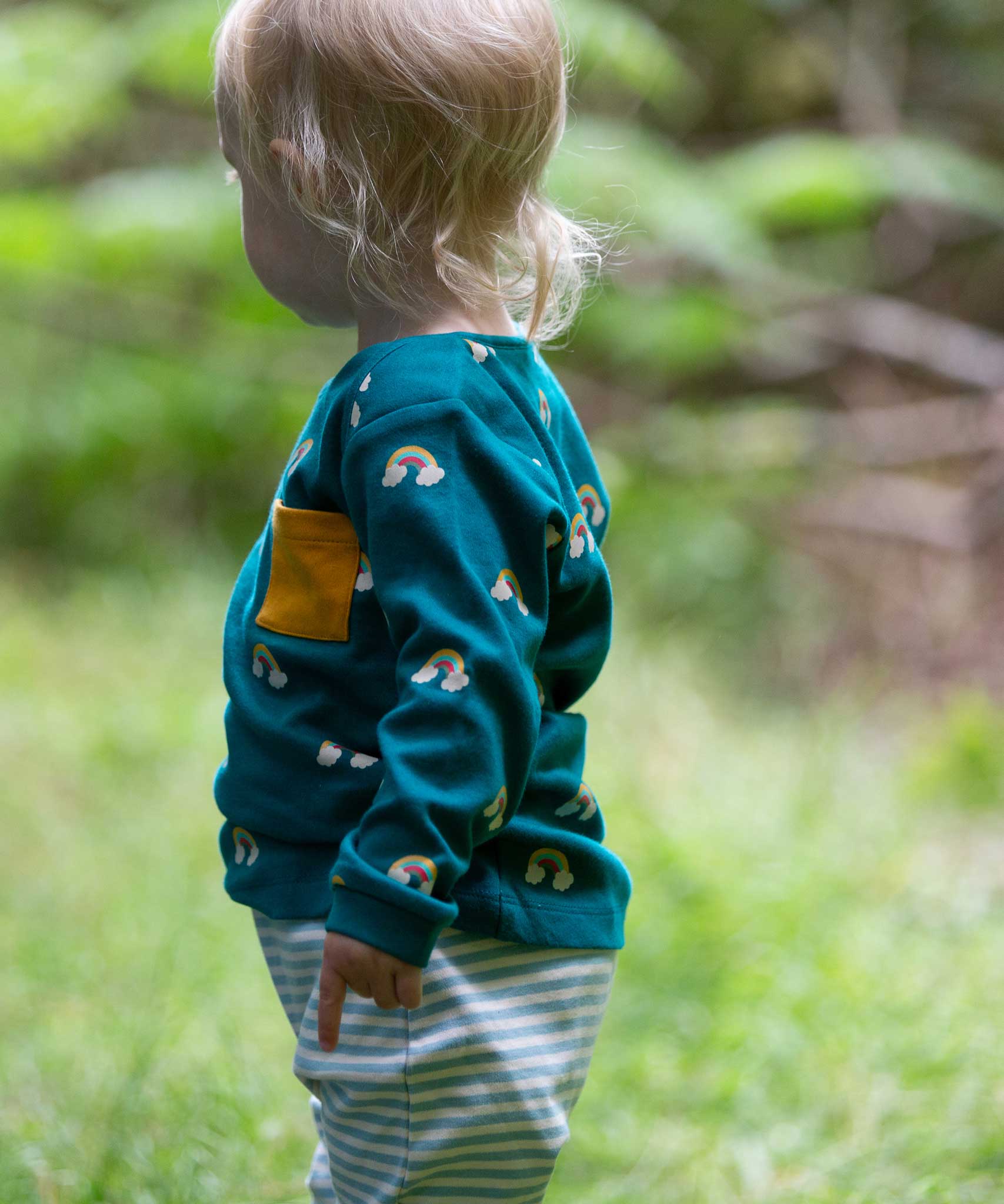 A side on view of a child wearing the Little Green Radicals Kids' Organic Top and Joggers Playset in the Turquoise Rainbows design. This play set is part of a wide range of organic cotton clothing for children available here at Babipur. 