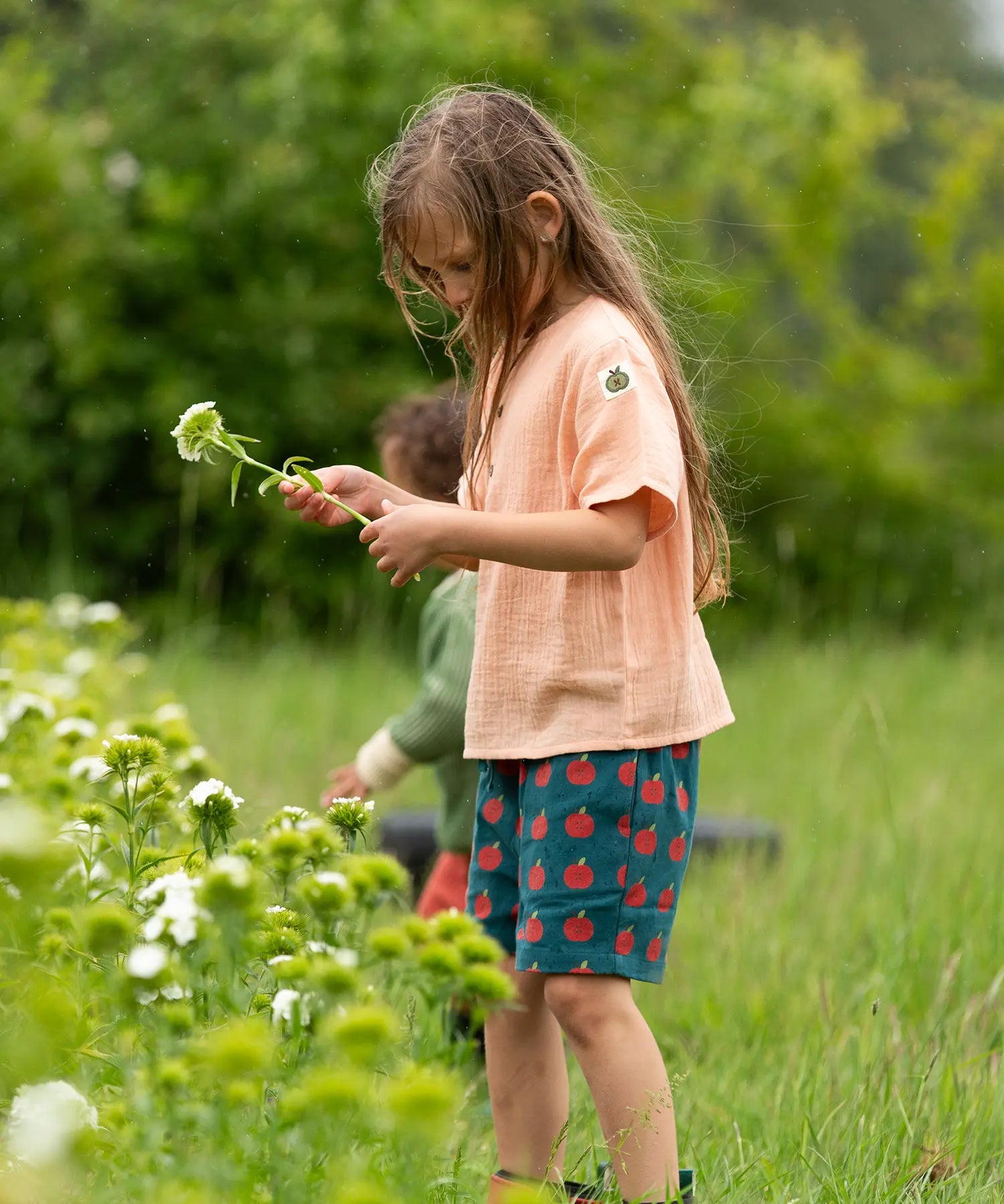 Little Green Radicals dark green blue kids organic cotton shorts with repeat apple print all worn by a child with a light orange t-shirt. 