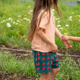 Little Green Radicals dark green blue kids organic cotton shorts with repeat apple print all worn by a child with a light orange t-shirt. 