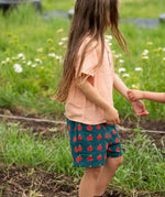 Little Green Radicals dark green blue kids organic cotton shorts with repeat apple print all worn by a child with a light orange t-shirt. 