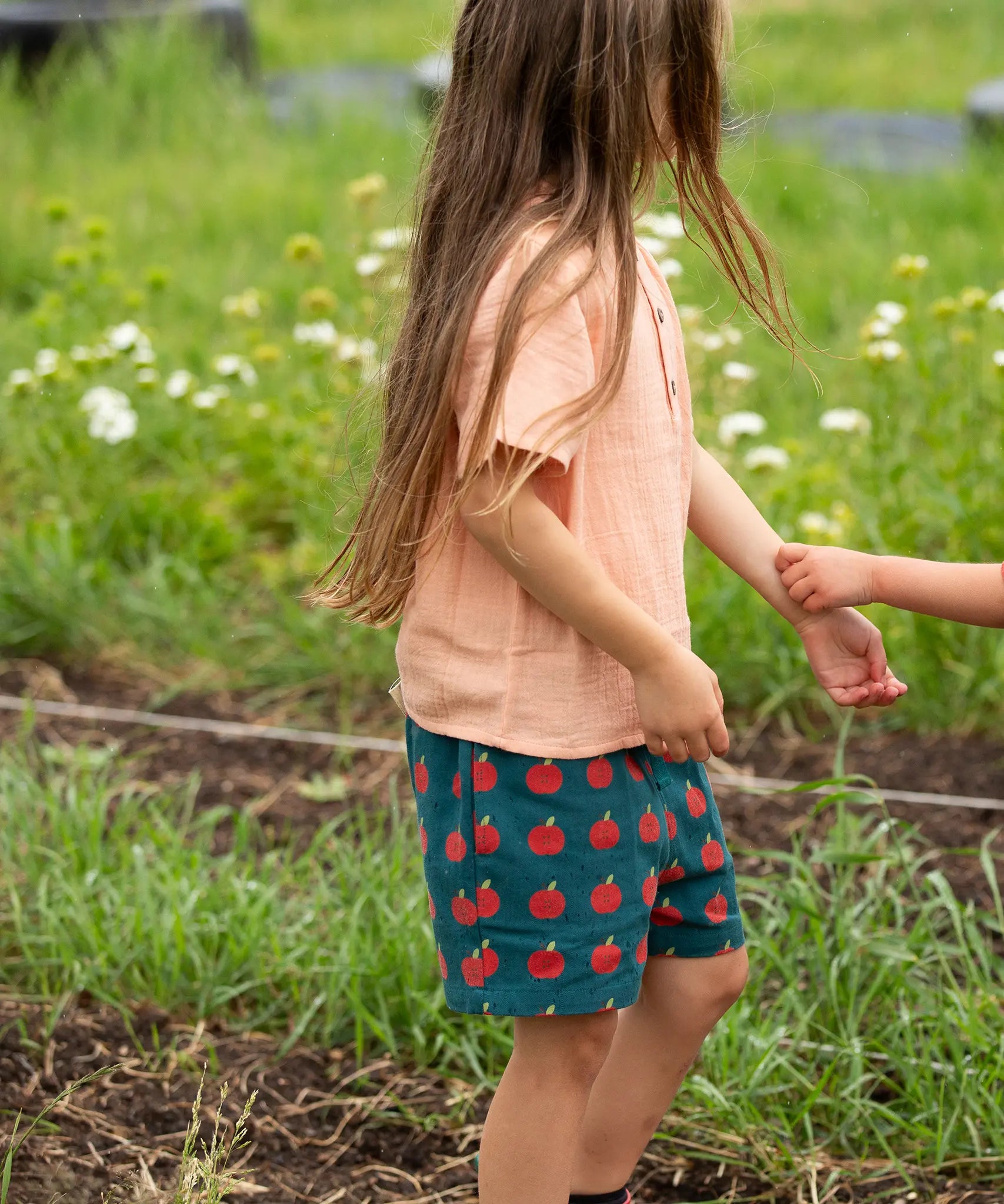 Little Green Radicals dark green blue kids organic cotton shorts with repeat apple print all worn by a child with a light orange t-shirt. 