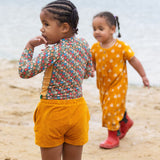 A child standing on a sandy beach wearing the Little Green Radicals Vintage Flowers design UPF 50+ Sun safe Long Sleeve Rash Vest with yellow gold shorts.