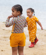 A child standing on a sandy beach wearing the Little Green Radicals Vintage Flowers design UPF 50+ Sun safe Long Sleeve Rash Vest with yellow gold shorts.
