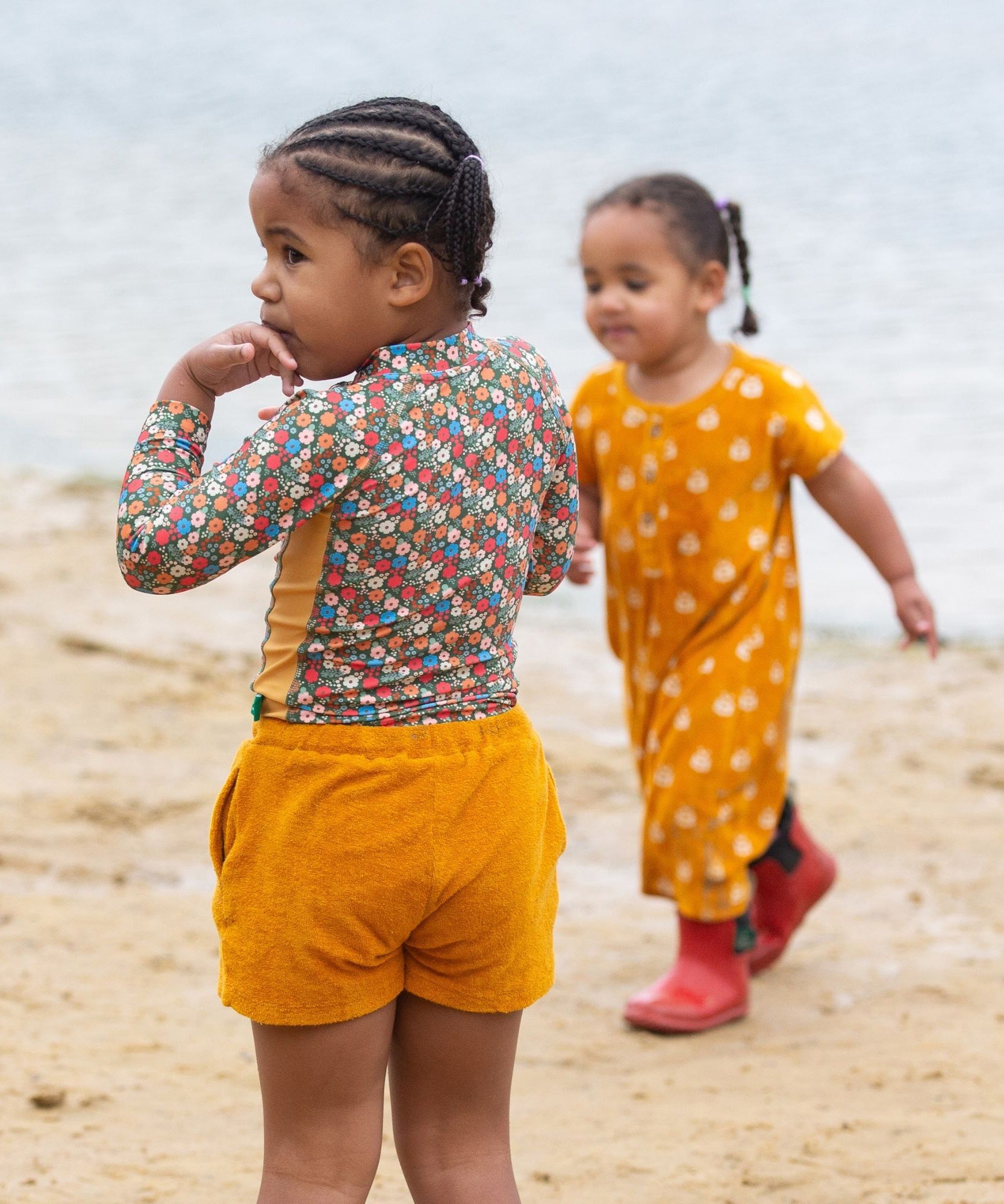 A child standing on a sandy beach wearing the Little Green Radicals Vintage Flowers design UPF 50+ Sun safe Long Sleeve Rash Vest with yellow gold shorts.