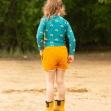A child wearing the Little Green Radicals Teal Rainbows UPF 50+ Sunsafe Long Sleeve Rash Vest with yellow gold shorts. The child is standing on the sand facing away from the camera showing the back of the vest.