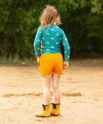 A child wearing the Little Green Radicals Teal Rainbows UPF 50+ Sunsafe Long Sleeve Rash Vest with yellow gold shorts. The child is standing on the sand facing away from the camera showing the back of the vest.