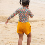 A child standing on a sandy beach wearing the Little Green Radicals Vintage Flowers design UPF 50+ Sun safe Long Sleeve Rash Vest with yellow gold shorts and yellow wellingtons. The child is facing away from the camera. 