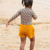 A child standing on a sandy beach wearing the Little Green Radicals Vintage Flowers design UPF 50+ Sun safe Long Sleeve Rash Vest with yellow gold shorts and yellow wellingtons. The child is facing away from the camera. 