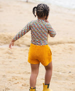 A child standing on a sandy beach wearing the Little Green Radicals Vintage Flowers design UPF 50+ Sun safe Long Sleeve Rash Vest with yellow gold shorts and yellow wellingtons. The child is facing away from the camera. 