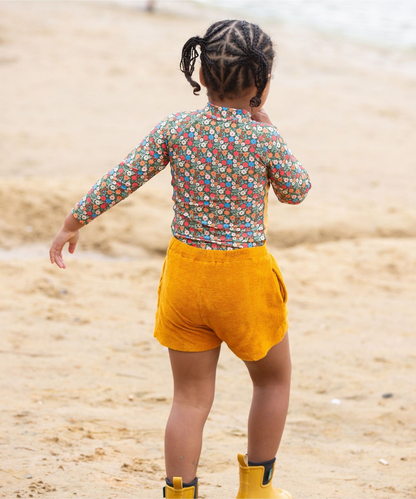 A child standing on a sandy beach wearing the Little Green Radicals Vintage Flowers design UPF 50+ Sun safe Long Sleeve Rash Vest with yellow gold shorts and yellow wellingtons. The child is facing away from the camera. 
