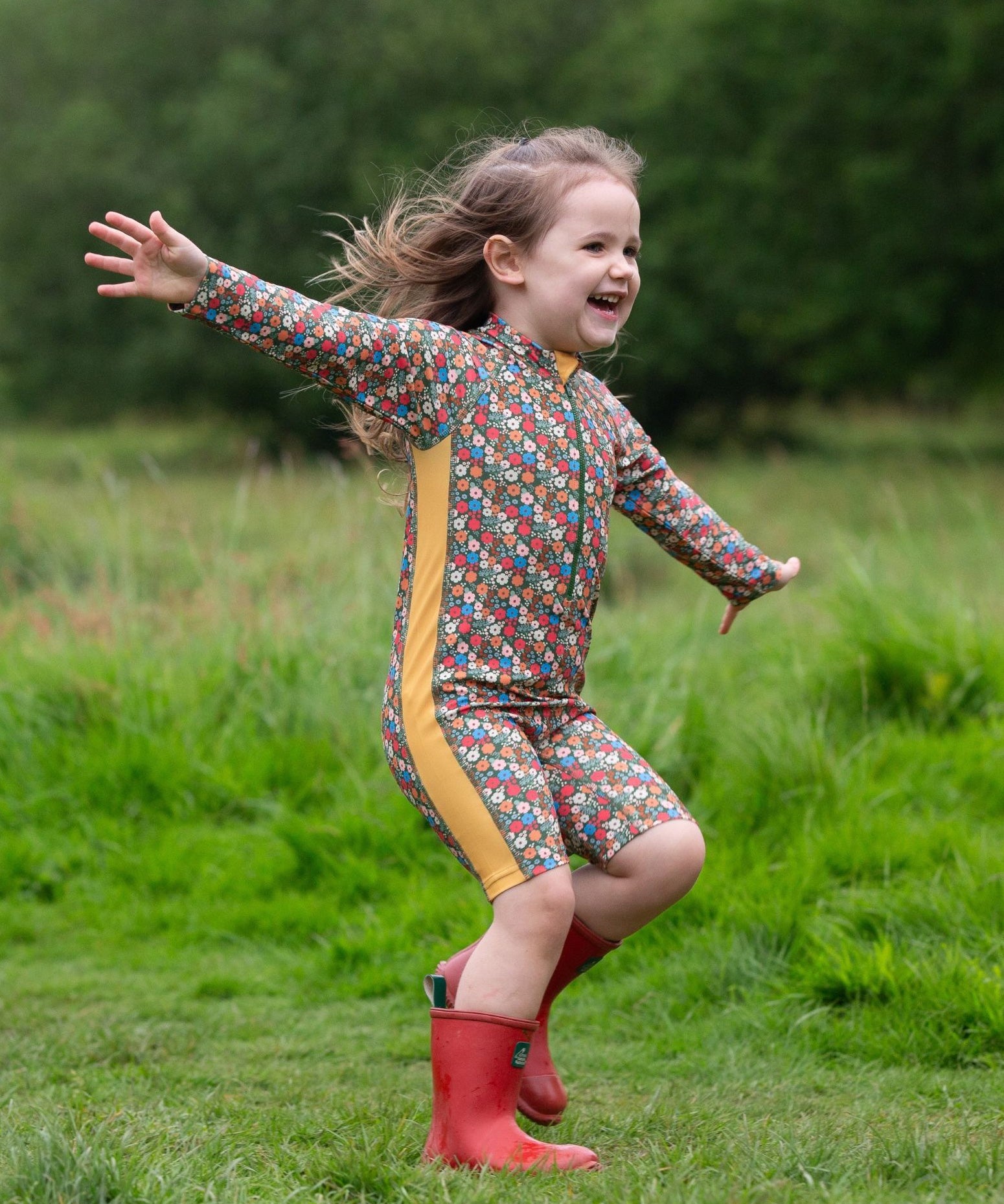A child wearing the Little Green Radicals Vintage Flowers UPF 50+ Recycled Sunsafe Sunsuit with a pair of red wellington boots.