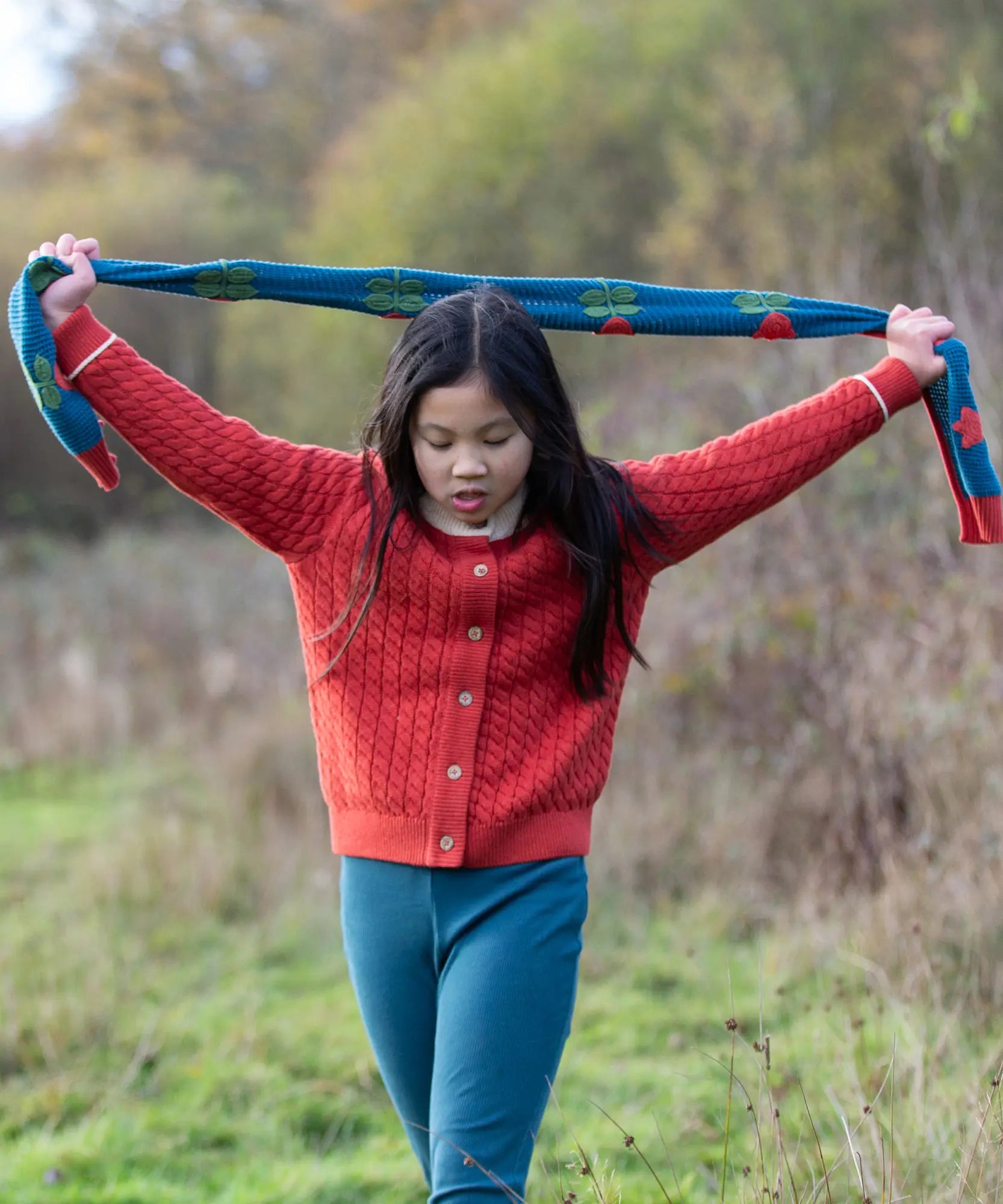 Child holding the Little Green Radicals tulips knitted blue scarf abover her head. Blue scarf with red tulips.