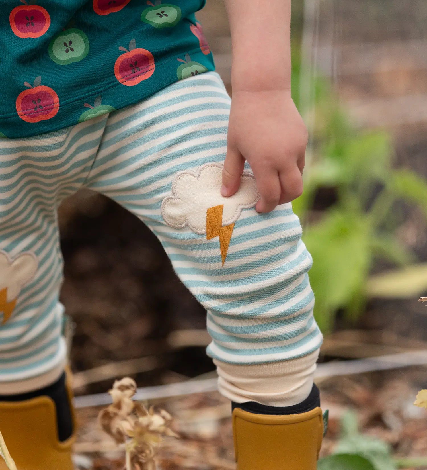 Close up of the Little Green Radicals weather knee patch blue and cream striped organic cotton joggers worn with a green apple print t-short and yellow wellington boots.