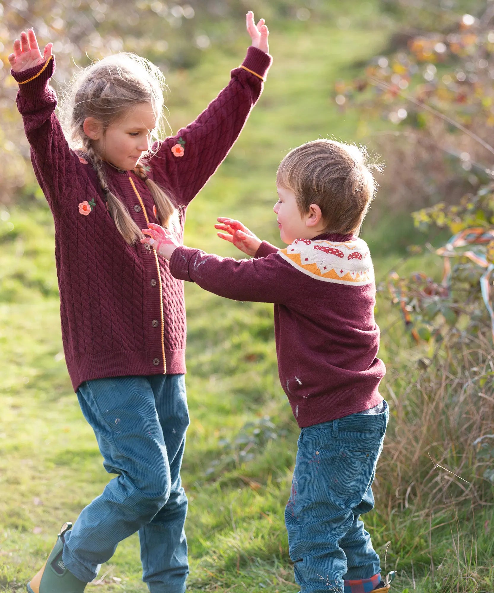 Child wearing the Little Green Radicals knitted organic cotton jumper with a mushroom Fair Isle design with green corduroy trousers.Another child is wearing a brown knitted cardigan withe corduroy trousers.