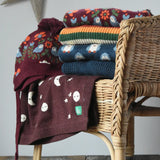 Stack of Little green Radicals knitted jumpers, knitted folk birds hat and moon blanket on a wicker chair. 