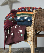 Stack of Little green Radicals knitted jumpers, knitted folk birds hat and moon blanket on a wicker chair. 