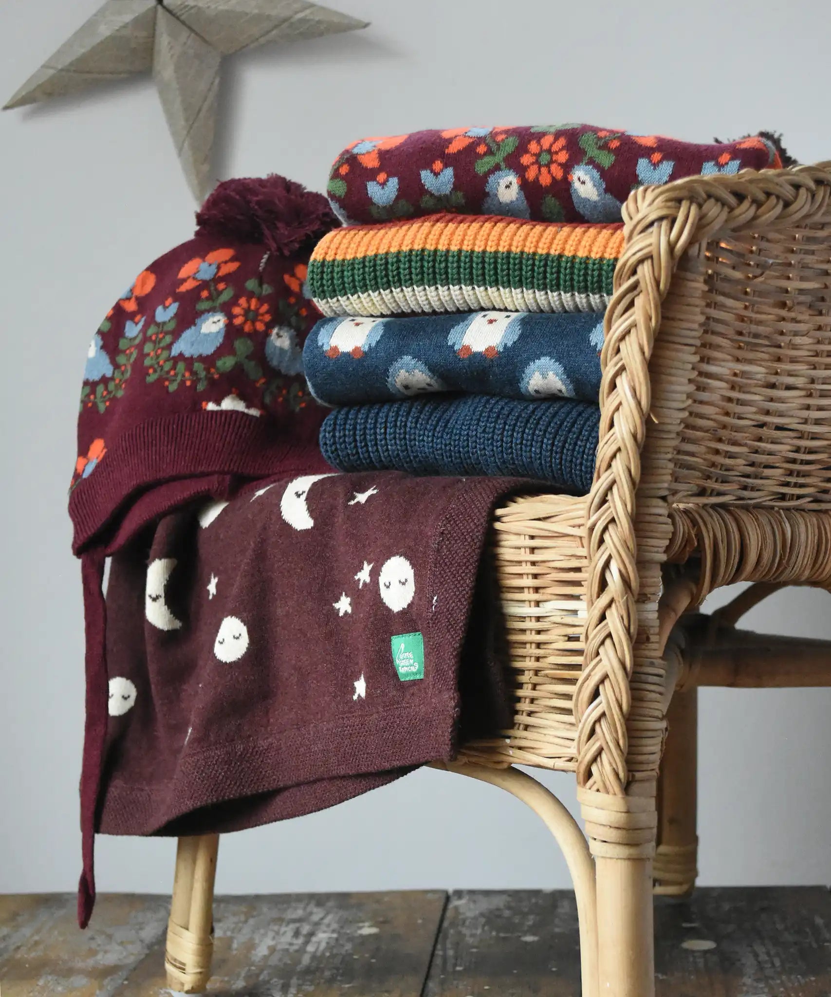 Stack of Little green Radicals knitted jumpers, knitted folk birds hat and moon blanket on a wicker chair. 