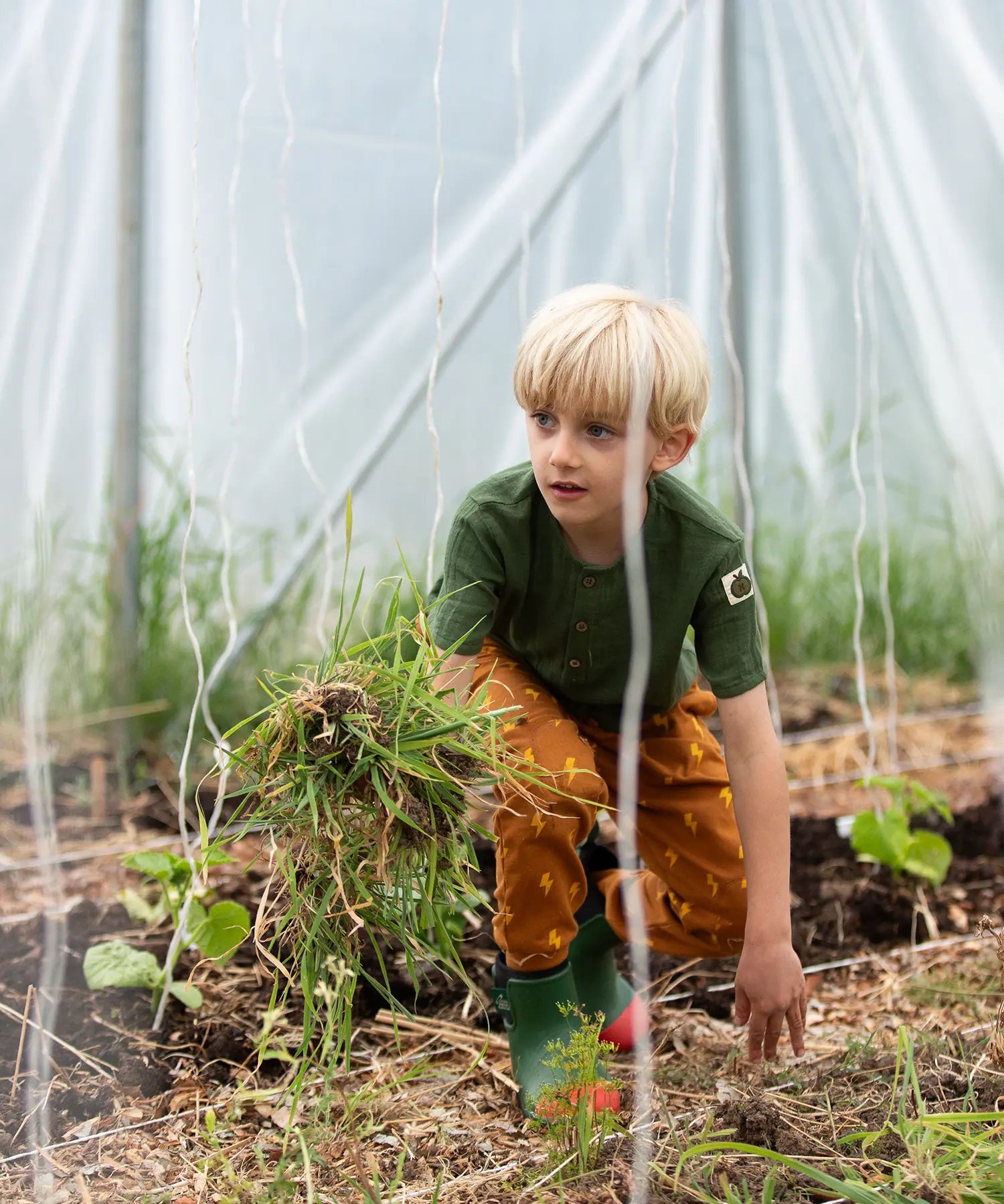 Child kneeling down wearing the Little Green Radicals yellow gold repeat lightning print cosy organic cotton kids joggers with a green henley muslin t-shirt and green and red wellington boots.