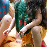 Close up of the Little Green Radicals kids dark green knitted sleeveless pullover with little cherry on the front being worn by a child with yellow shorts.