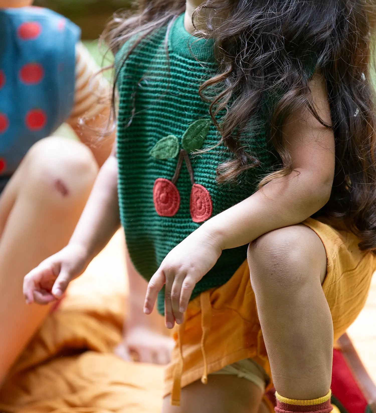 Close up of the Little Green Radicals kids dark green knitted sleeveless pullover with little cherry on the front being worn by a child with yellow shorts.
