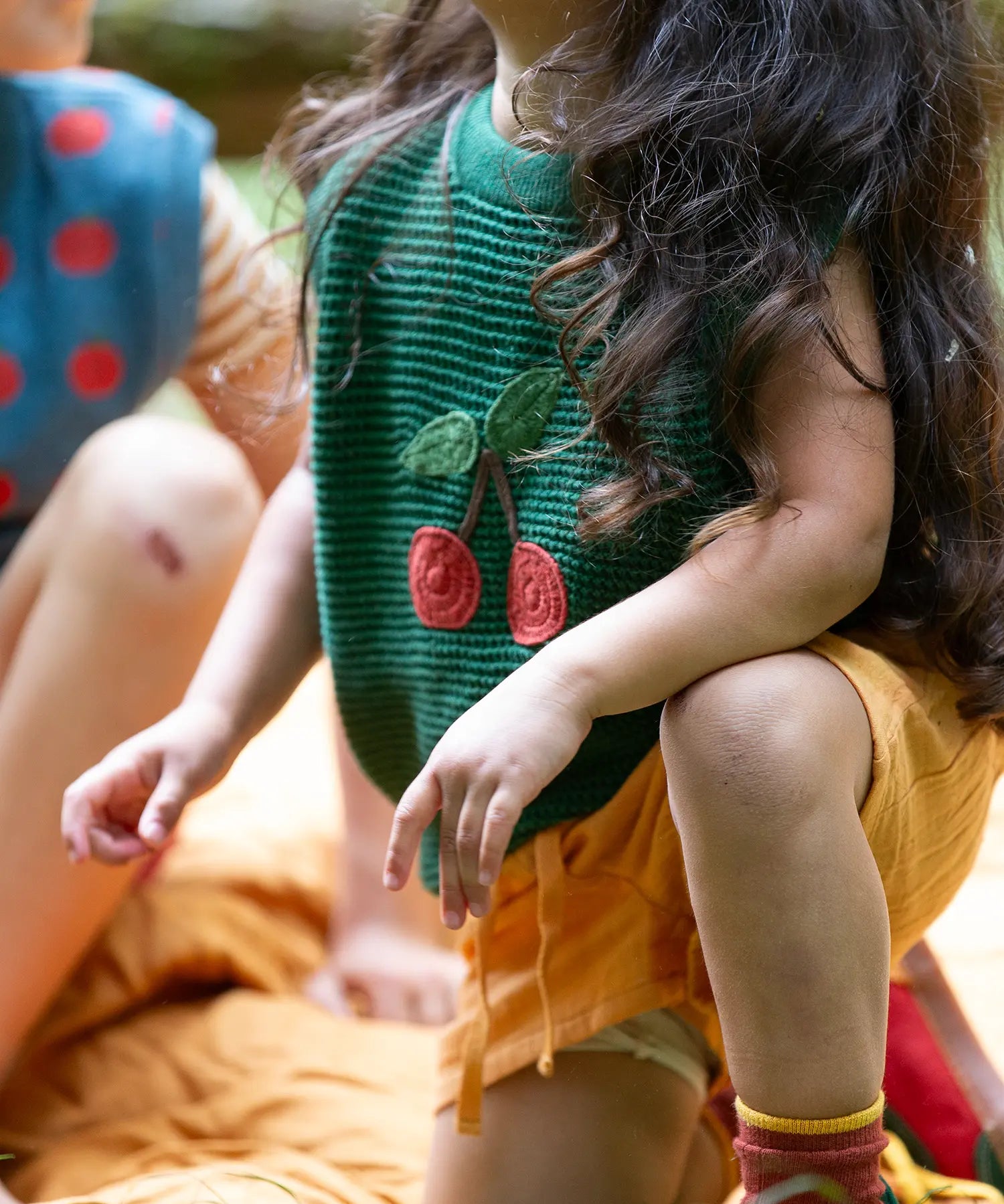 Close up of the Little Green Radicals kids dark green knitted sleeveless pullover with little cherry on the front being worn by a child with yellow shorts.