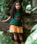 Child wearing the Little Green Radicals kids dark green knitted sleeveless pullover with little cherry on the front with golden ochre yellow shorts.