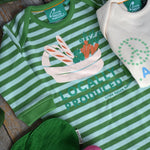 Little Green Radicals Locally Produced Playsuit placed on a wooden surface surrounded by different vegetables. An Oli & Carol Ramona The Radish Mini Doudou Baby Teether is placed on the playsuit 