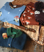 Collection of Children's clothing draped on a wicker hamper. Maxomorra owl top, mushroom knitted joggers, moon knitted hat and blue knitted cardigan. 