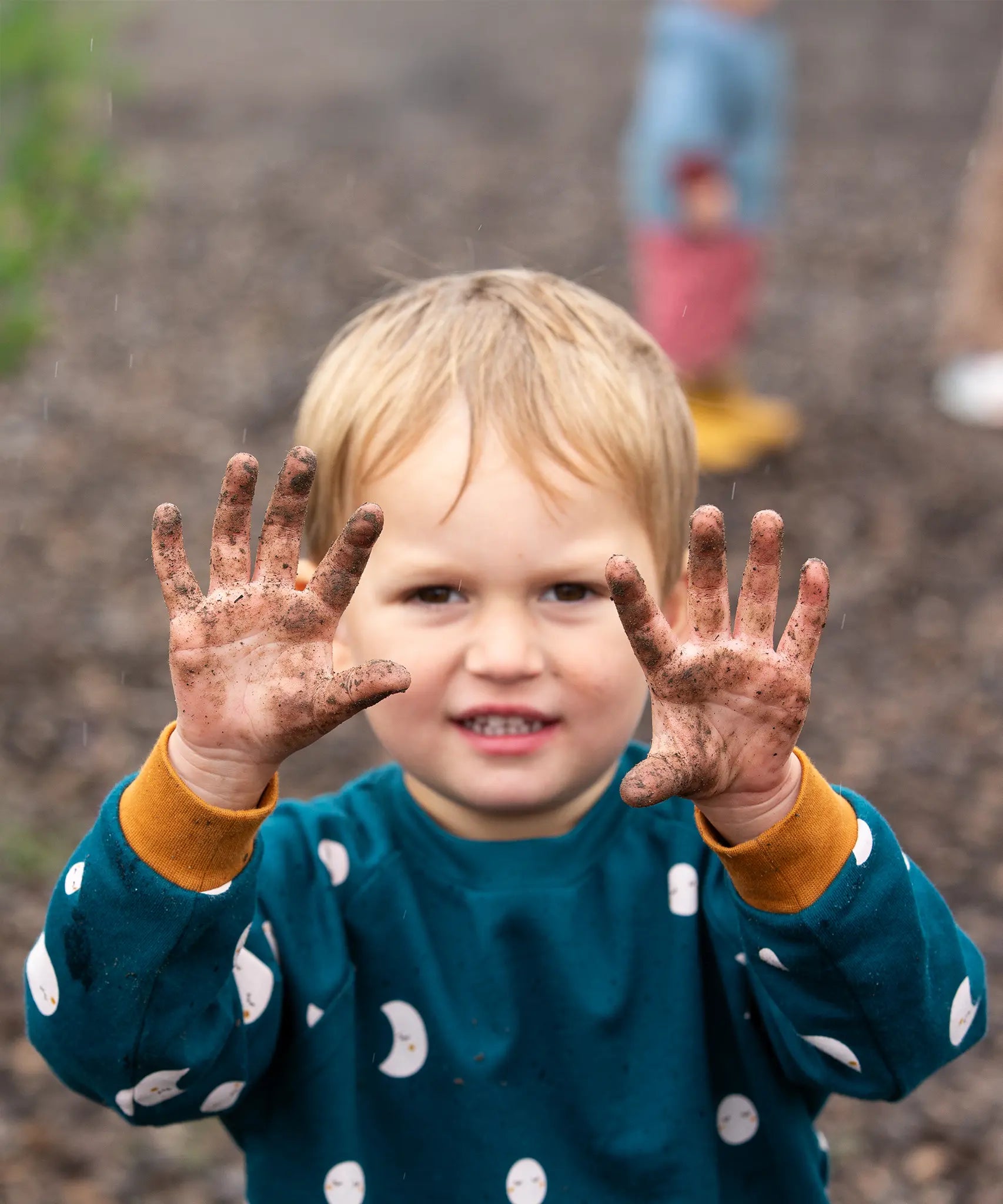 Close up of child wearing the Little Green Radicals midnight moon print blue raglan organic-cotton children's sweatshirt holding his soil covered hands up to the camera.