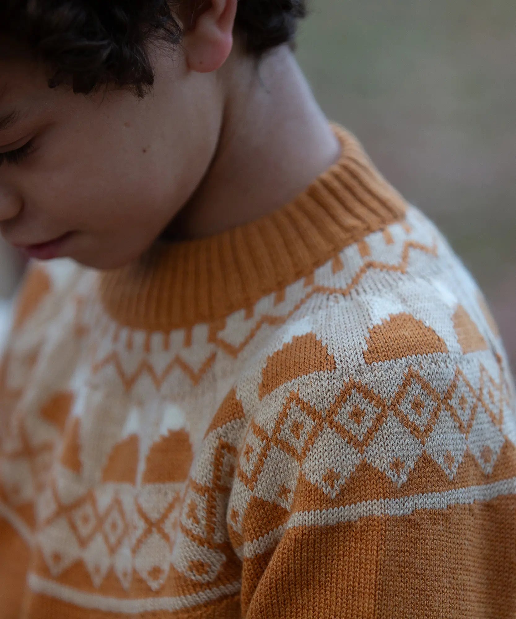 Close up of a child wearing the Little Green Radicals mountain Fair Isle snuggly woollen jumper in yellow gold. The Fair Isle details n around the neckline featuring snow topped mountains surrounded by patterns.