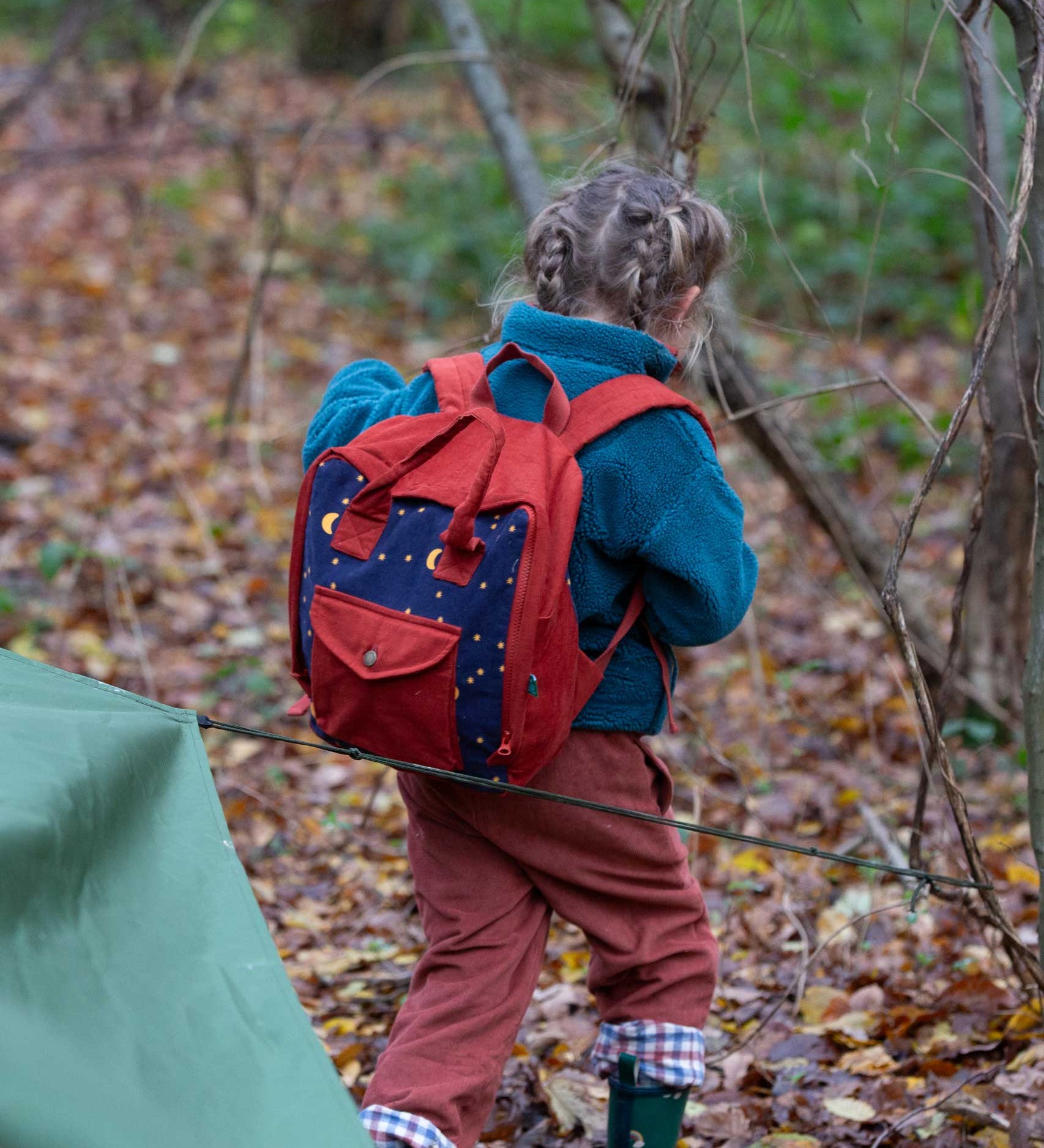 A child walking through a forest wearing the Little Green Radicals dark nights corduroy backpack on their back. A 100% organic cotton backpack in a navy and red corduroy style with a starry night design. Features a handle on the top for easy carrying a front pocket and 2 bottle holders. This children's bag is a part of a wide range of organic cotton clothing and accessories for children available here at Babipur. 