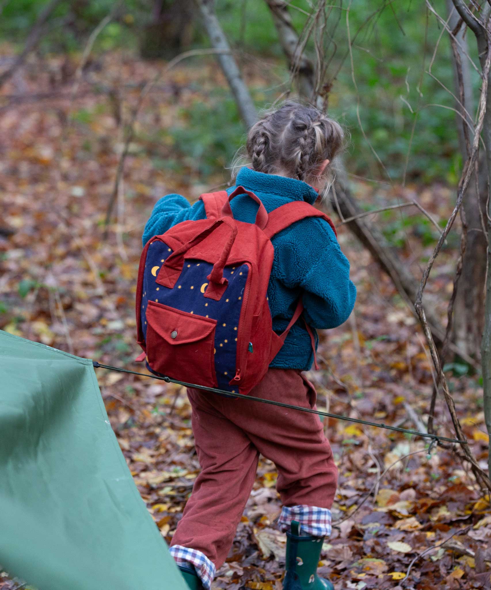 A child walking through a forest wearing the Little Green Radicals dark nights corduroy backpack on their back. A 100% organic cotton backpack in a navy and red corduroy style with a starry night design. Features a handle on the top for easy carrying a front pocket and 2 bottle holders. This children's bag is a part of a wide range of organic cotton clothing and accessories for children available here at Babipur. 