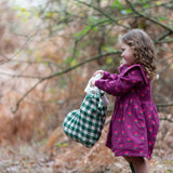 Child holding a Little Green Radicals  olive green check organic cotton stocking wearing a purple dress and green wellington boots.