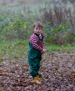 Child wearing a Little Green Radicals olive green recycled lined waterproof dungarees with a striped top underneath and yellow gold ankle wellingtons.
