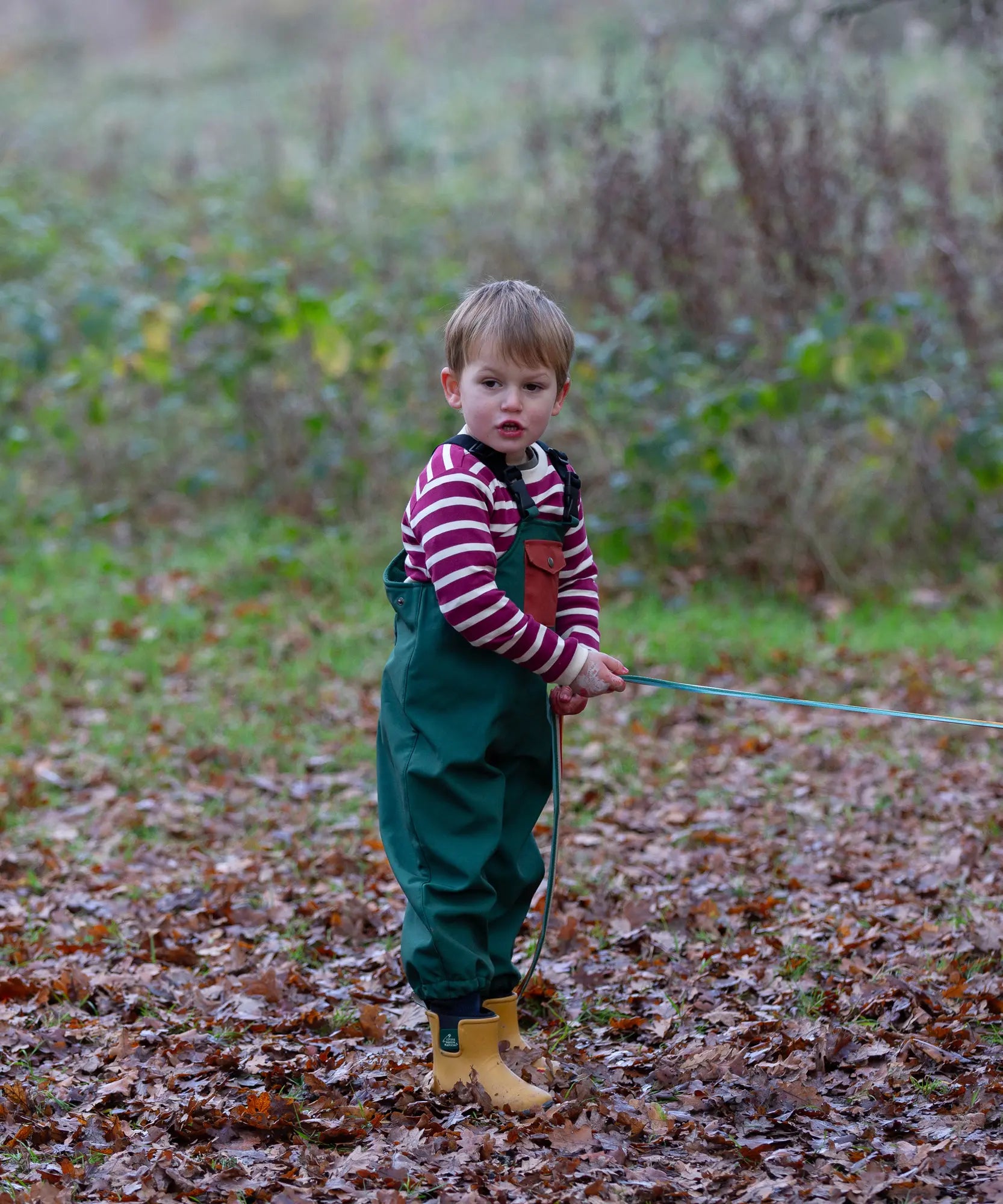Child wearing a Little Green Radicals olive green recycled lined waterproof dungarees with a striped top underneath and yellow gold ankle wellingtons.