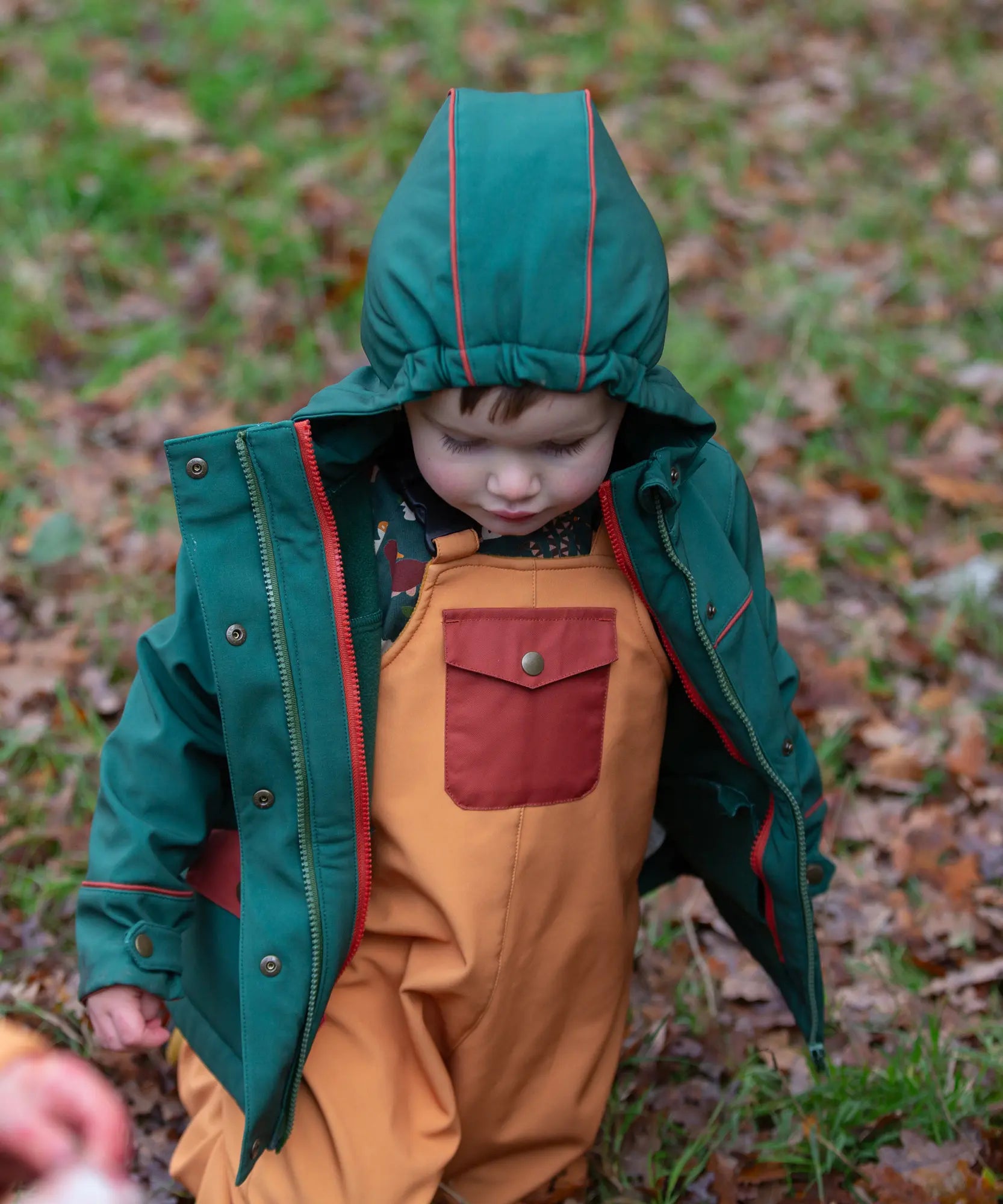 Child wearing a Little Green Radicals olive green recycled waterproof 3 in 1 coat with yellow waterproof dungarees underneath.