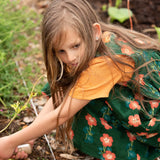 Little Green Radicals green orange blooms repeat print reversible pinny kids dress being worn with a yellow t-short underneath and red wellington boots.