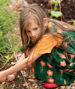 Little Green Radicals green orange blooms repeat print reversible pinny kids dress being worn with a yellow t-short underneath and red wellington boots.
