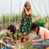 Little Green Radicals green orange blooms repeat print reversible pinny kids dress being worn with a yellow t-short underneath and red wellington boots.