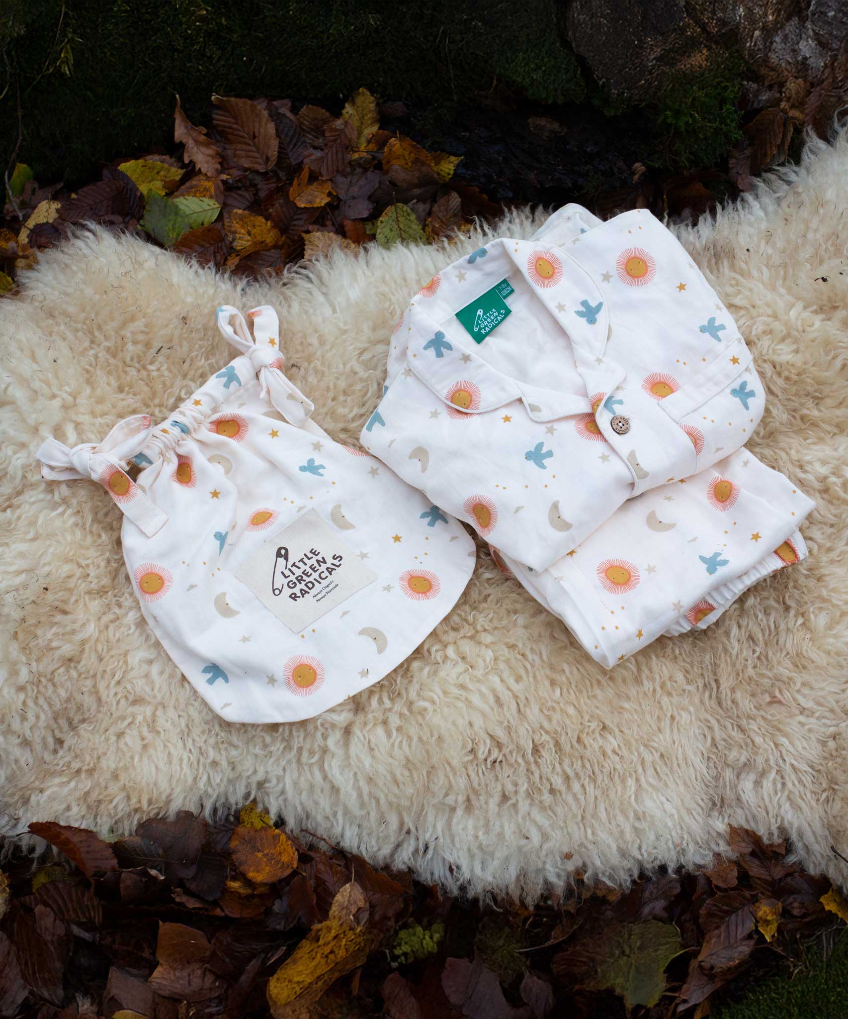 Little Green Radicals moon and sun classic pyjama set shown folded and placed on a sheepskin rug. A GOTS organic cotton pyjama set including a long sleeve shirt style top in light pink with prints of cute birds flying through stars the moon and sun also includes matching bottoms. These pyjamas are part of a wide range of organic cotton clothing for children available here at Babipur.