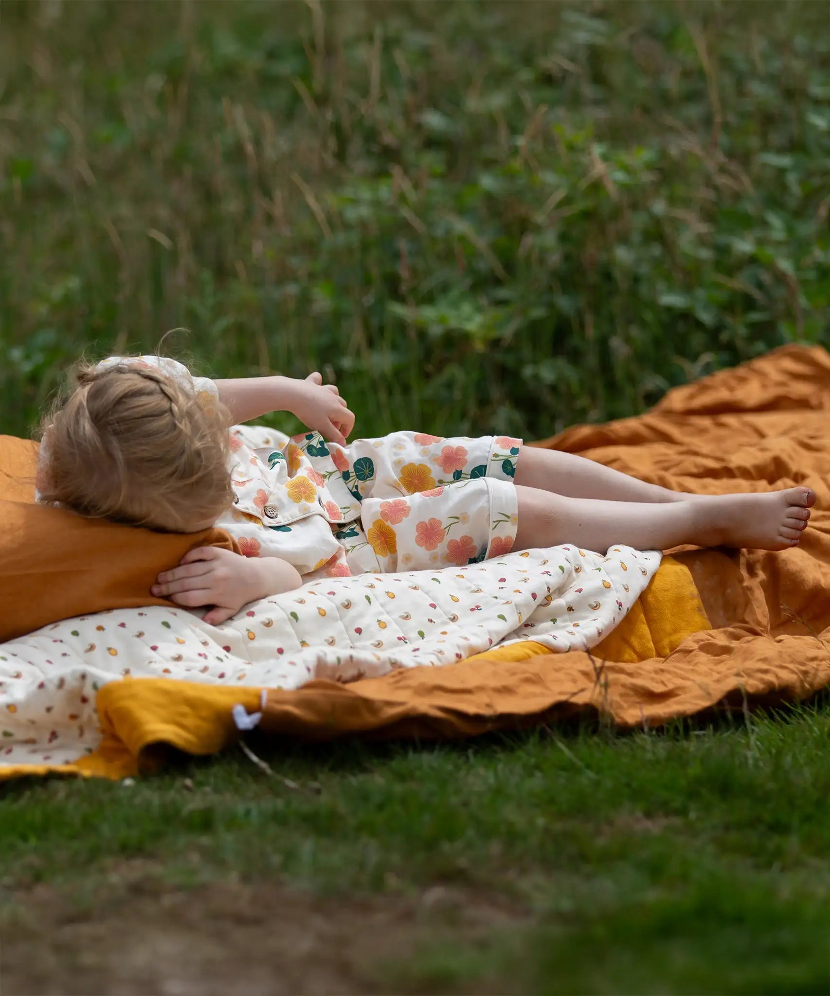 Child laying on a blanket outdoors wearing the Little Green Radicals organic cotton short pyjamas set in cream with a pink and yellow flora print. 