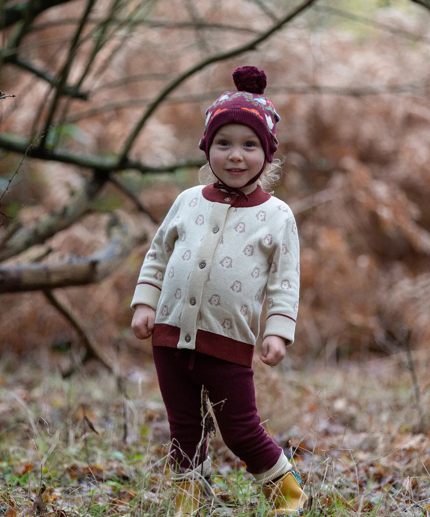 Child wearing the Little Green Radicals organic cotton knitted cardigan with snow penguins design, maroon trousers and a folk birds knitted bobble hat.
