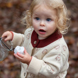 Child wearing the Little Green Radicals organic cotton knitted cardigan with snow penguins design. Natural cream cardigan with penguins all over, maroon trim on the collar and hem line.