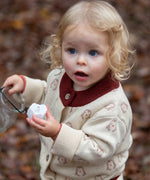 Child wearing the Little Green Radicals organic cotton knitted cardigan with snow penguins design. Natural cream cardigan with penguins all over, maroon trim on the collar and hem line.
