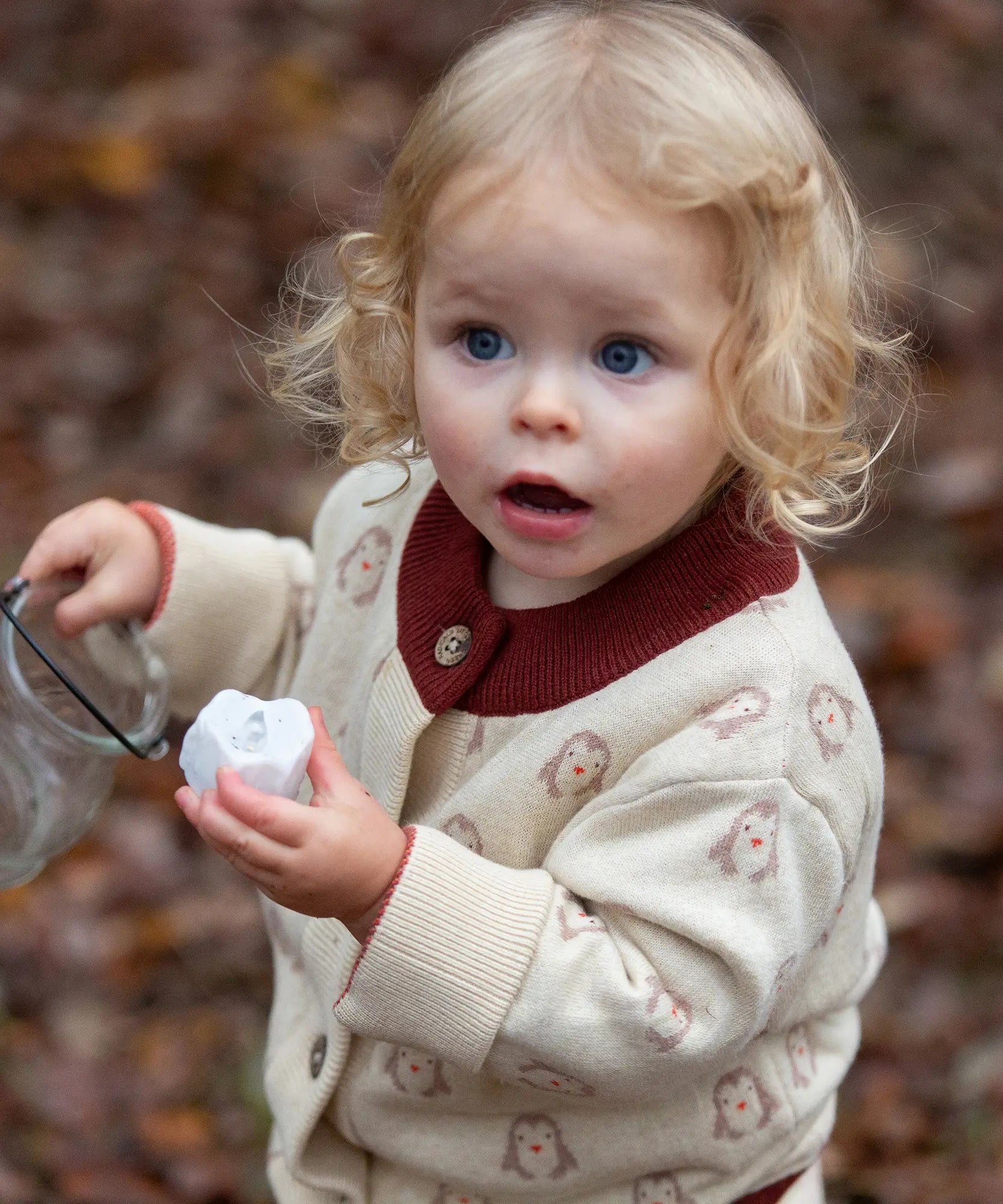 Child wearing the Little Green Radicals organic cotton knitted cardigan with snow penguins design. Natural cream cardigan with penguins all over, maroon trim on the collar and hem line.