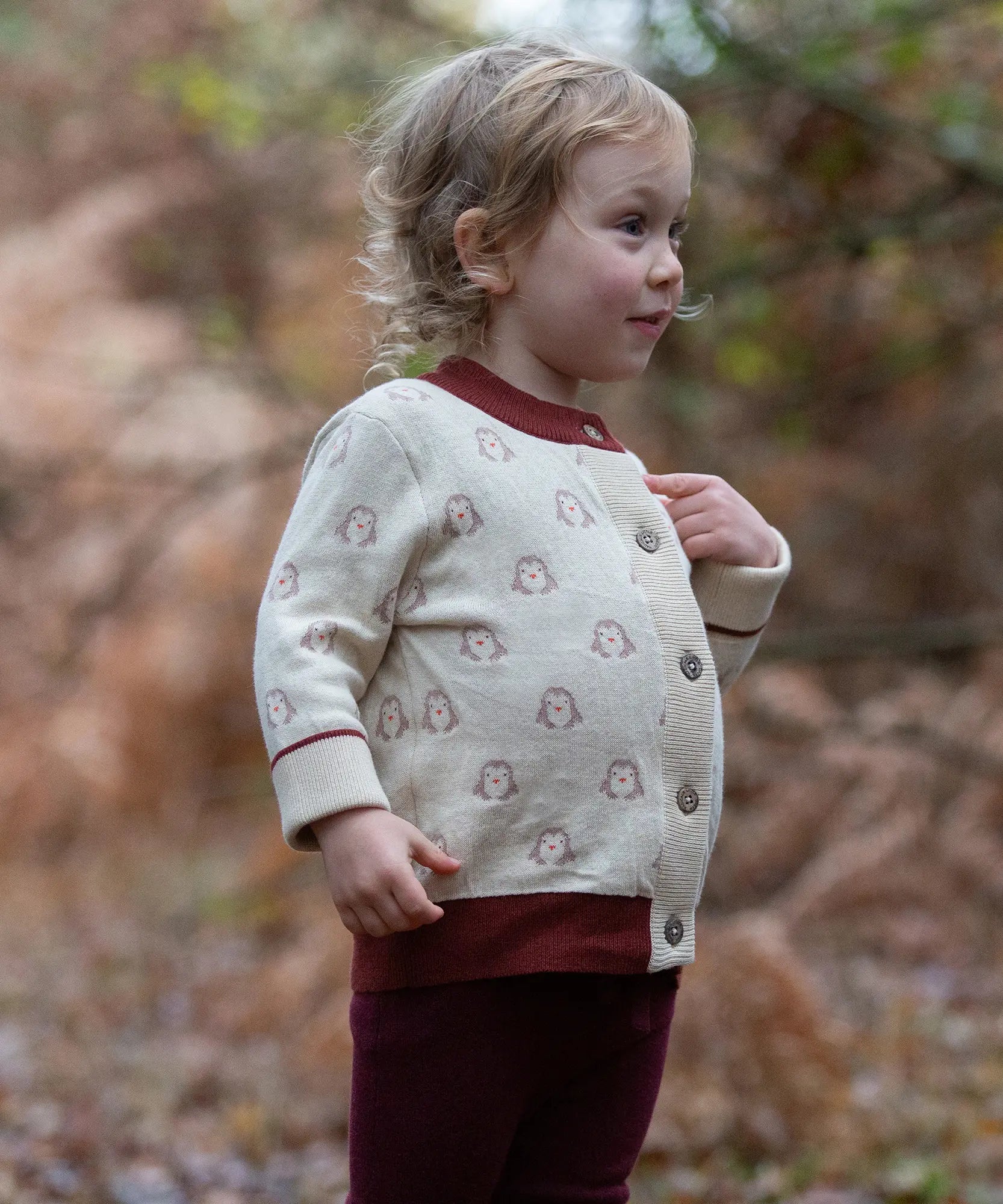 Child wearing the Little Green Radicals organic cotton knitted cardigan with snow penguins design. Natural cream cardigan with penguins all over, maroon trim on the collar and hem line.