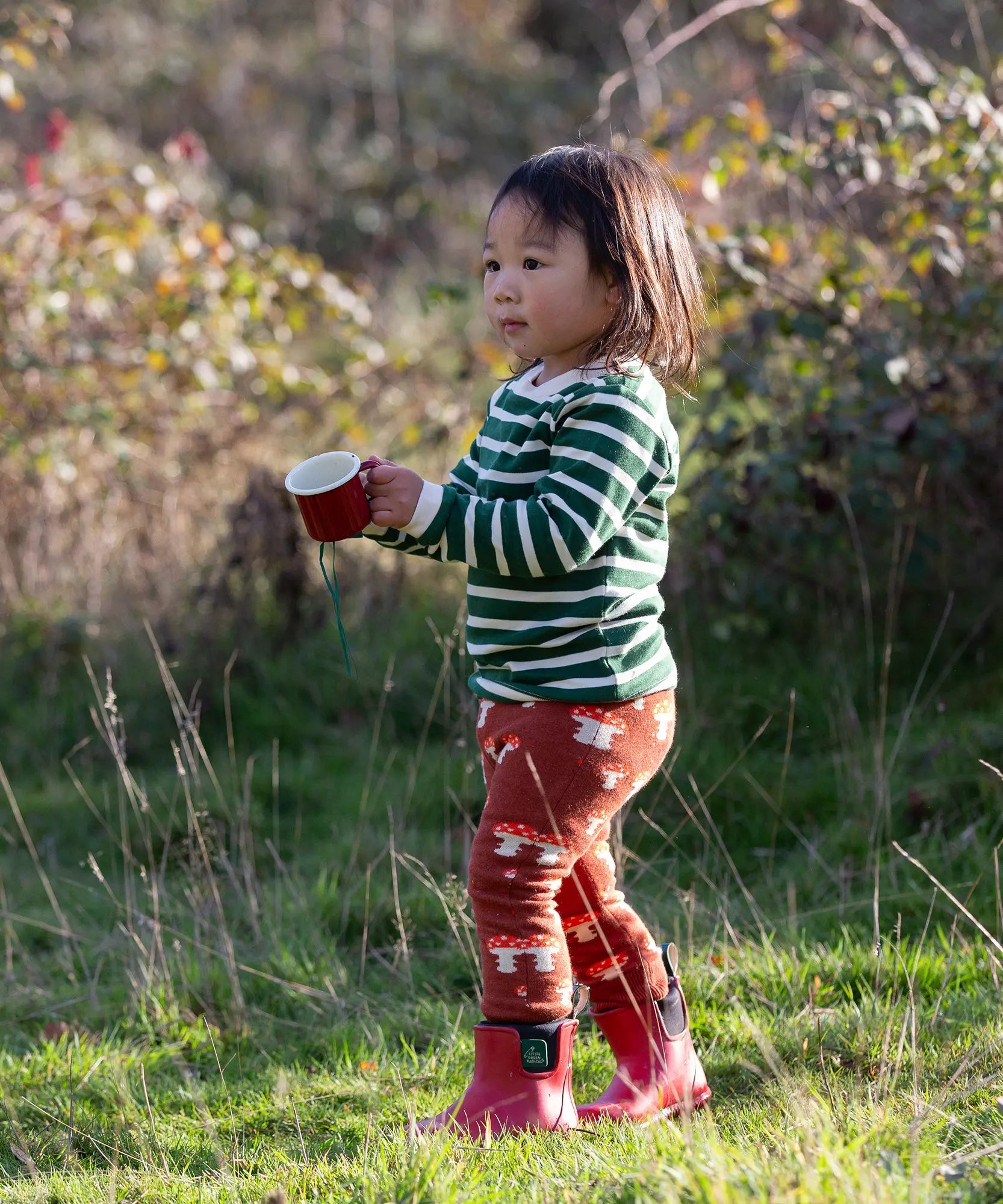 Child wearing the Little Green Radicals organic cotton knitted joggers with a mushroom design with red wellingtons and green striped long sleeved top.