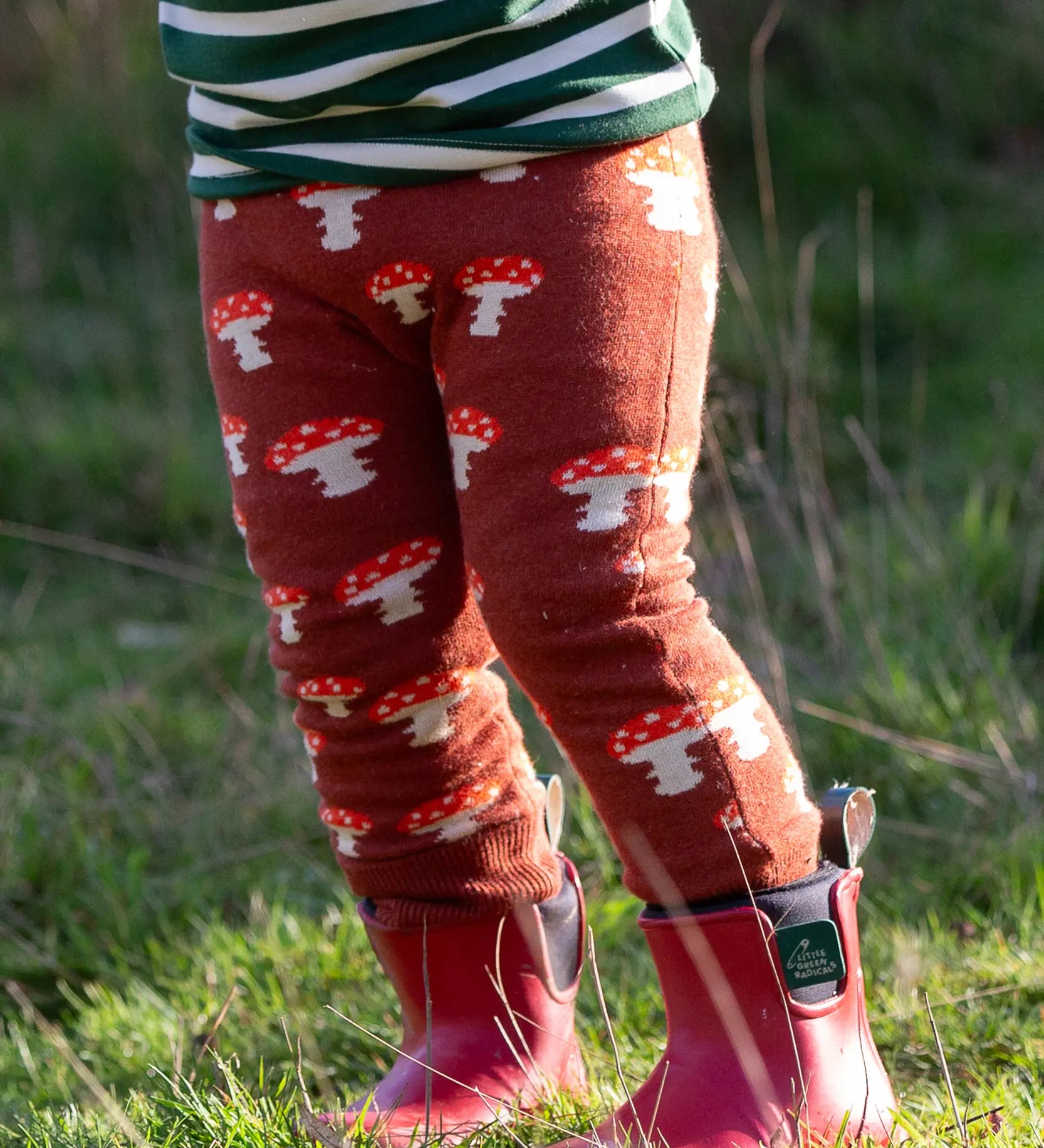 Child wearing the Little Green Radicals organic cotton knitted joggers with a mushroom design with red wellingtons. Rust orange knitted leggings featured red and white mushrooms.