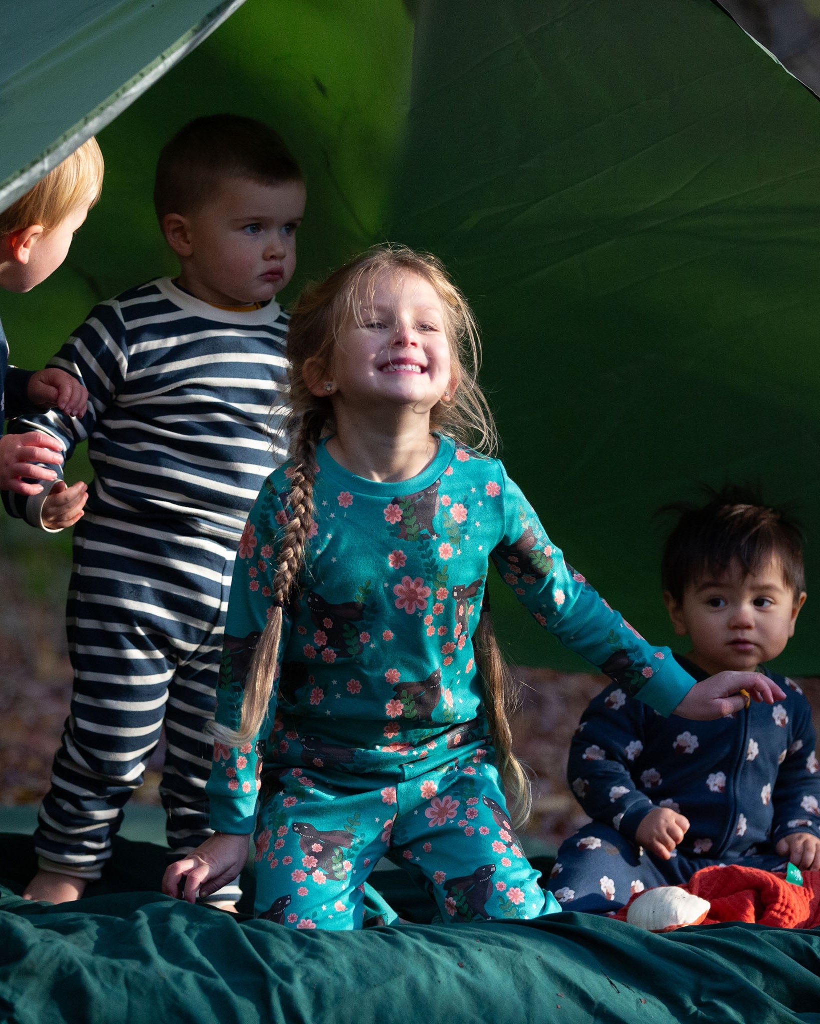 Children standing outside a tent in the forest. They are all wearing Little Green Radicals organic ccotton pyjamas. One is wearing a pair with the Winter Rabbit on them, one in wearing navy and white striped design and the third is wearing the sleeping sheep print. Little green Radicals have a wide range of organic cotton, Fairtrade children's clothing an outerwear available here at Babipur.